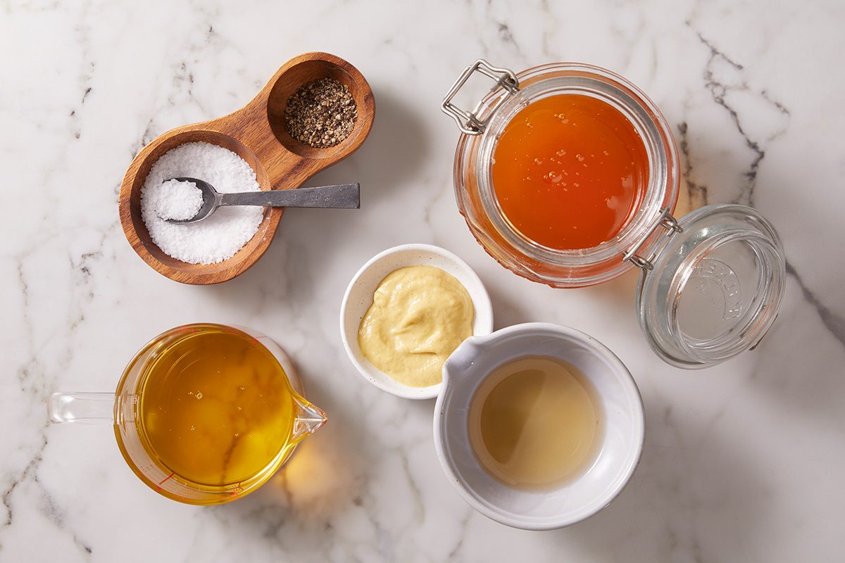 A marble surface displays small bowls of salt and pepper, a jar of honey, a cup of oil, a bowl of mustard, and a cup of vinegar, arranged neatly for a recipe.