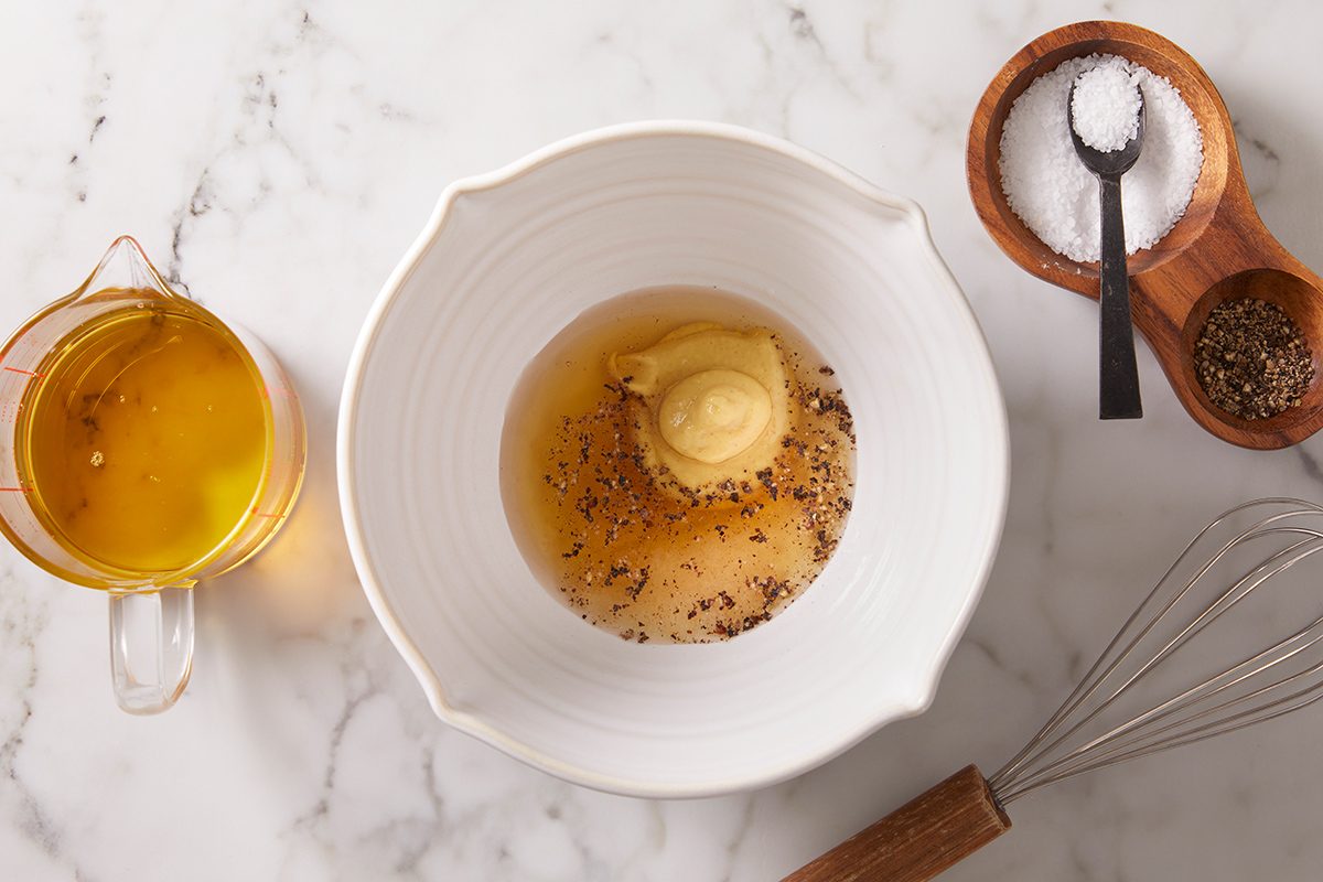 A white bowl with mustard, spices, and vinegar sits on a marble surface. Nearby are a wooden salt and pepper holder with a spoon, a glass cup of oil, and a whisk.