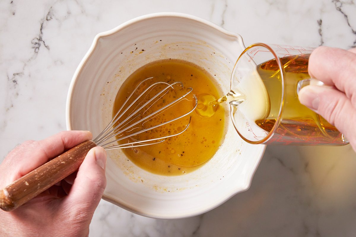 A person whisks a golden liquid in a white bowl while slowly pouring oil from a glass, preparing a dressing or vinaigrette on a marble countertop.
