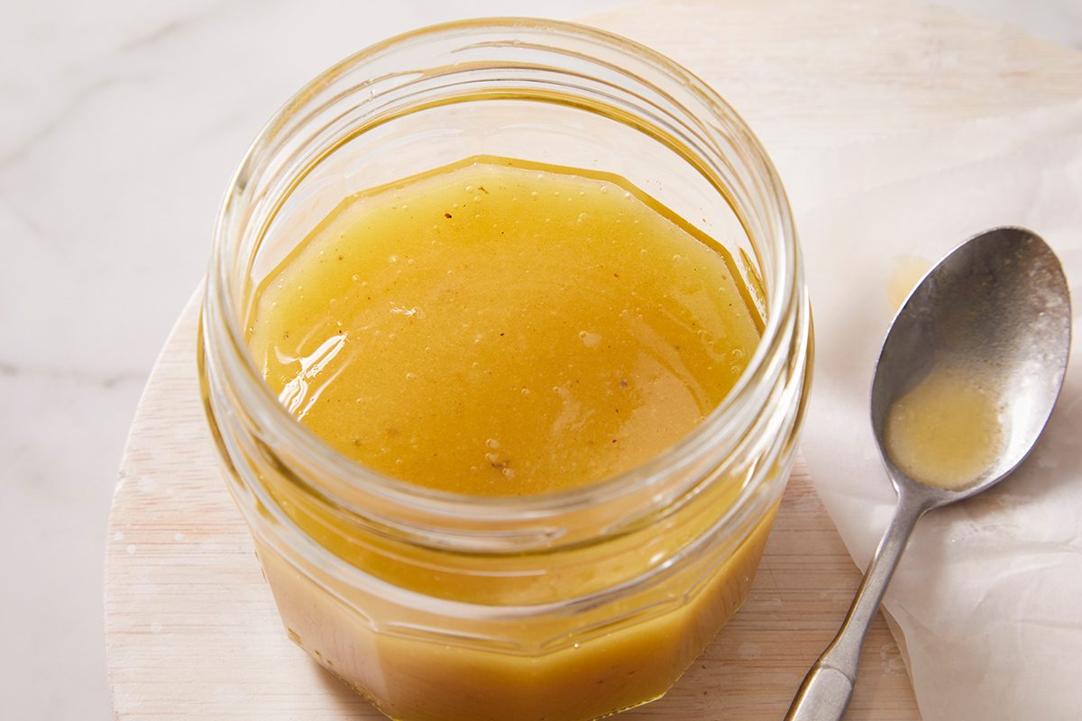 An open glass jar filled with golden yellow salad dressing sits on a light surface next to a metal spoon with some dressing on it.