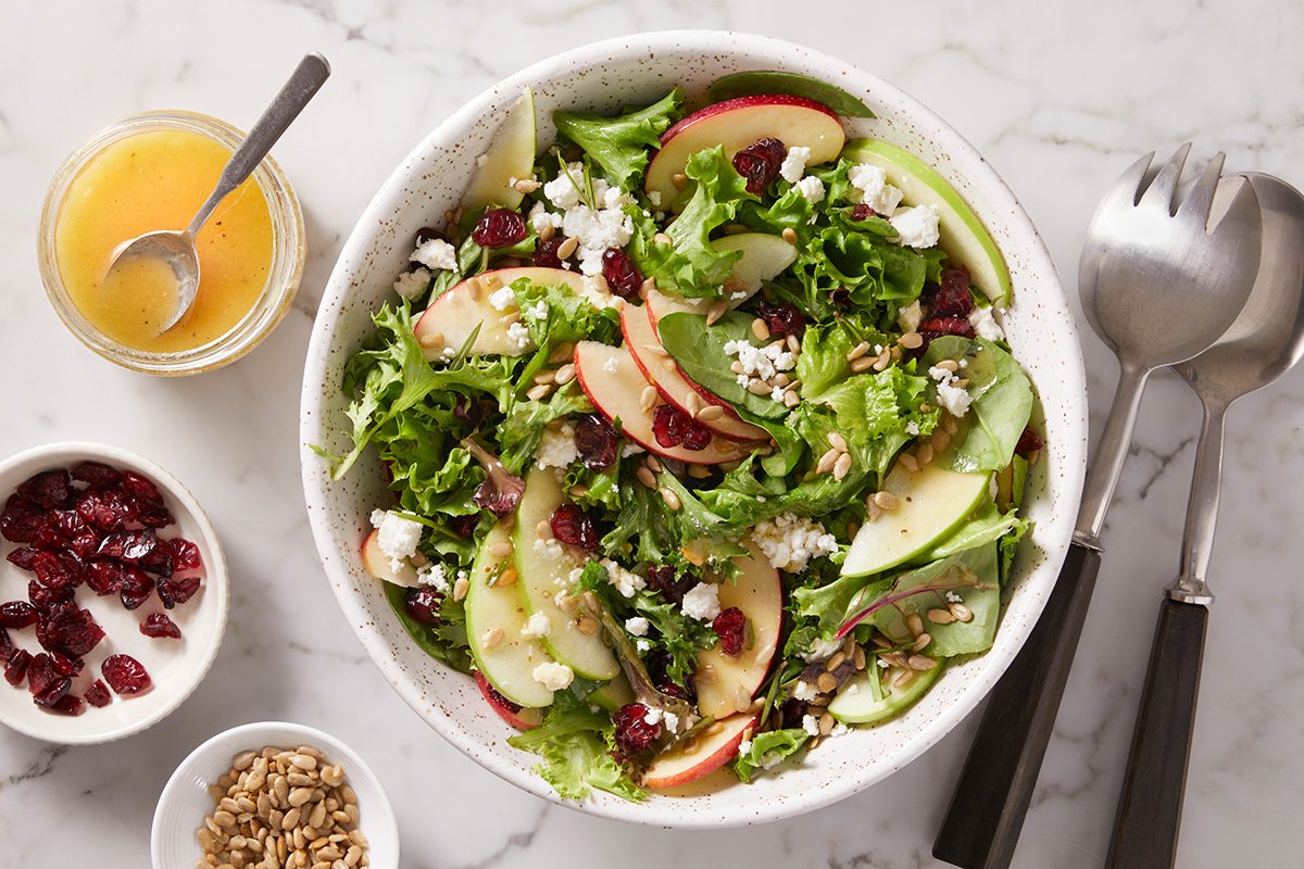 A bowl of mixed green salad with sliced apples, avocado, dried cranberries, and crumbled cheese. Nearby are small bowls of dressing, dried cranberries, and nuts, with serving utensils beside the salad.