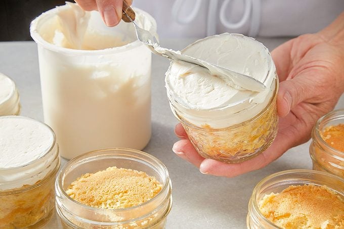 Eye Level horizontal image of a hand using an offset spatula to spread white frosting from a large tub onto a dessert in a small glass jar. Several other partially prepared desserts in glass jars are visible on a light-colored surface.