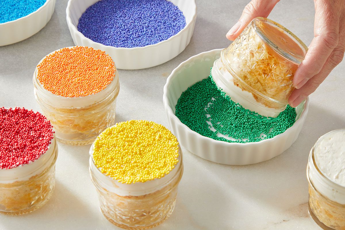 Eye Level horizontal image of a hand dipping a dessert in a glass jar into a white fluted ramekin filled with green sprinkles. Several other glass jar desserts are visible on a light surface, some already topped with red, orange, or yellow sprinkles. Additional ramekins in the background hold blue and purple sprinkles, showcasing the decorating process.