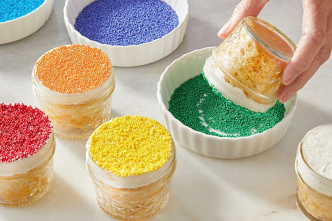 Eye Level horizontal image of a hand dipping a dessert in a glass jar into a white fluted ramekin filled with green sprinkles. Several other glass jar desserts are visible on a light surface, some already topped with red, orange, or yellow sprinkles. Additional ramekins in the background hold blue and purple sprinkles, showcasing the decorating process.