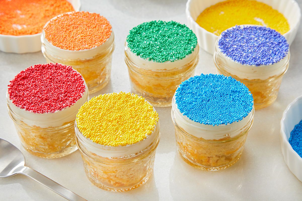 Eye Level horizontal image of six individual desserts in glass jars, each topped with white frosting and vibrant colorful sprinkles. The sprinkles are red, orange, yellow, green, blue, and purple, with small white dishes containing additional sprinkles and a spoon visible nearby
