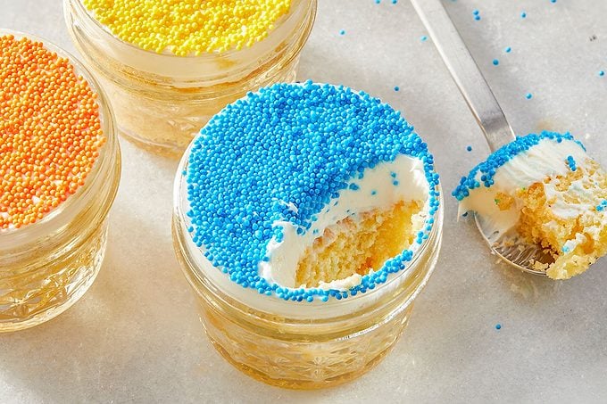 Eye level horizontal image of three individual cake jars on a white marble surface. One jar, topped with blue sprinkles, has a spoonful taken out, revealing the cake and frosting, with the spoon resting beside it. Two other jars with orange and yellow sprinkles are partially visible in the background