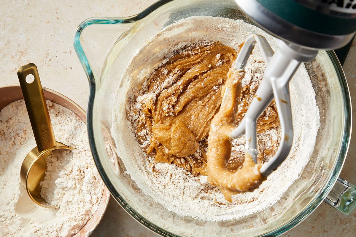 Overhead horizontal image of a KitchenAid mixer combining wet and dry ingredients in a clear mixing bowl. There is flour in the bowl and in a measuring cup to the left. | [Original] RMS 281760 Iced Chestnut Cookies with Bourbon Brown Butter Icing; How to; Step 2 Add remaining browned butter and brown sugar to a large mixing bowl; beat until well combined. Add chestnut spread; egg yolks; bourbon and vanilla bean paste. Combine the flour; cinnamon; baking soda; cream of tartar and salt; gradually add to brown sugar mixture and mix well. (Show bowl of dry ingredients and half of it being mixing into wet ing bowl)