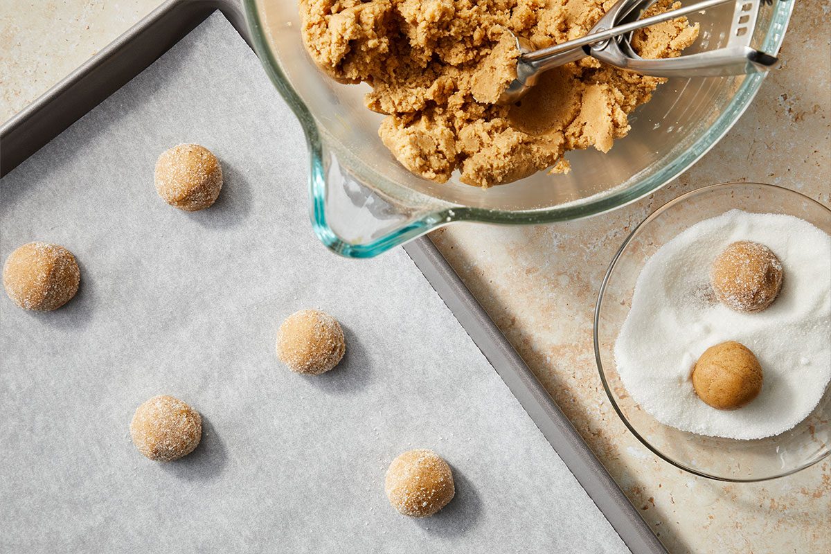 Overhead horizontal image of cookie dough balls being prepared. Cookie dough is shown in a clear glass mixing bowl, along with a cookie scoop, while some dough balls are displayed rolled in sugar and some dough balls are placed on a baking sheet. | [Original] RMS 281760 Iced Chestnut Cookies with Bourbon Brown Butter Icing; How to; Step 3 Place sugar in a small bowl. Shape dough into 1-in. balls; roll in sugar. Place 2 in. apart on parchment-lined baking sheets. (self explanatory)