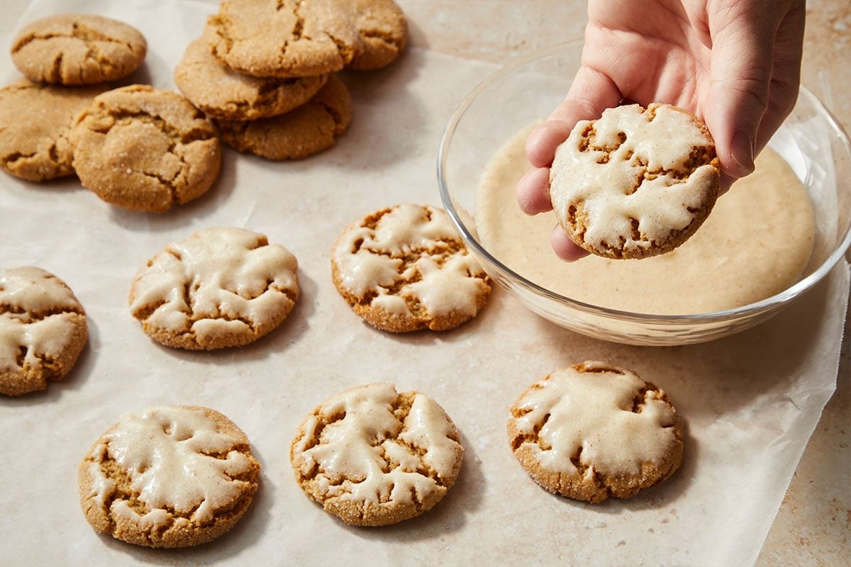 Eye level horizontal image of glazed cookies. A hand holds a glazed cookie over a glass bowl of glaze while other glazed and unglazed cookies sit on parchment paper. | [Original] RMS 281760 Iced Chestnut Cookies with Bourbon Brown Butter Icing; How to; Step 4 In a medium bowl combine reserved 1/2 cup browned butter; confectioners' sugar; milk; bourbon; vanilla paste and salt. Dip each cookie top halfway into icing; allow excess to drip off. Place on waxed paper; let stand until set. (self explanatory)