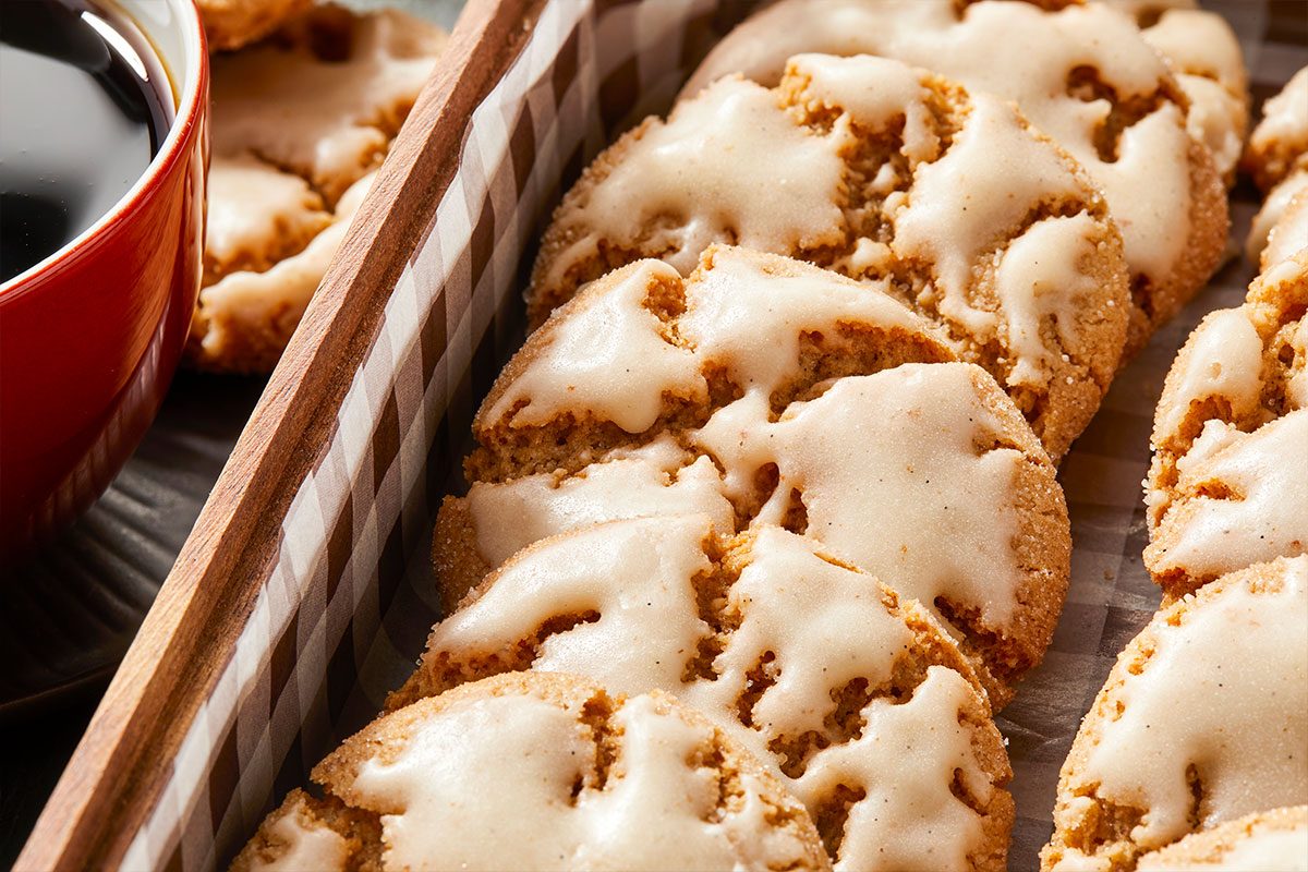 Eye Level horizontal image of iced cookies in a wooden tray next to a red mug filled with black coffee. The cookies are golden brown and covered with a thin layer of sweet white icing. | [Original] RMS 281760 Iced Chestnut Cookies with Bourbon Brown Butter Icing; Horizontal; Pull