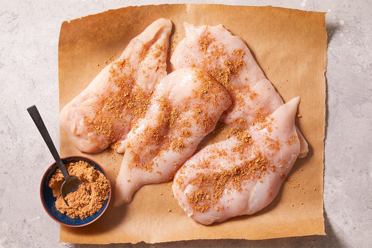 Overhead horizontal image of four raw chicken breasts sprinkled with seasonings. A bowl filled with the same seasoning mix sits to the side with a spoon. The chicken sits on a piece of parchment paper.