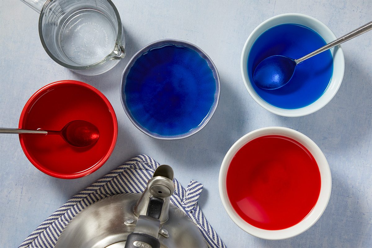 Overhead horizontal image of four bowls containing red and blue liquids, with spoons resting in two of the bowls. A glass measuring cup filled with clear liquid, a silver kettle, and a blue and white striped kitchen towel are also visible on a light background.