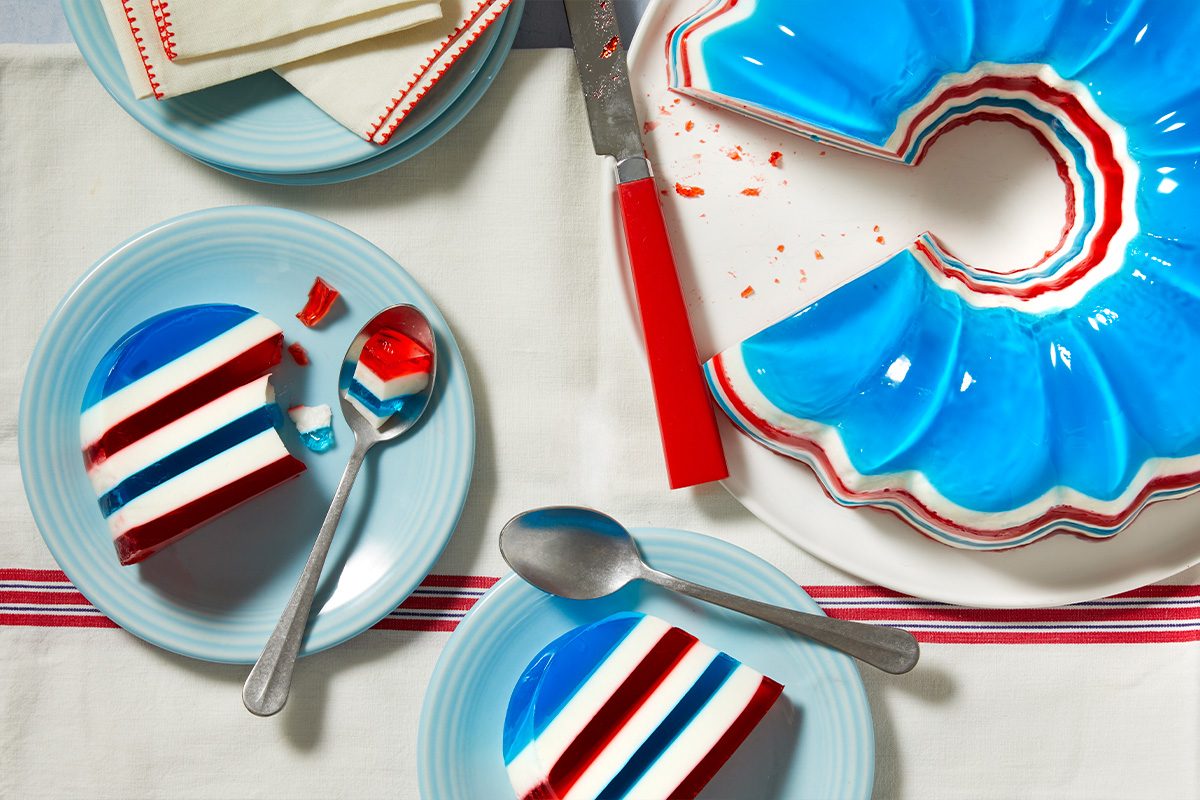 Overhead horizontal image of a festive red, white, and blue layered gelatin dessert. A large fluted gelatin mold sits on a white plate, with several slices served on light blue plates alongside spoons. The dessert, napkins, and tablecloth evoke a patriotic theme.