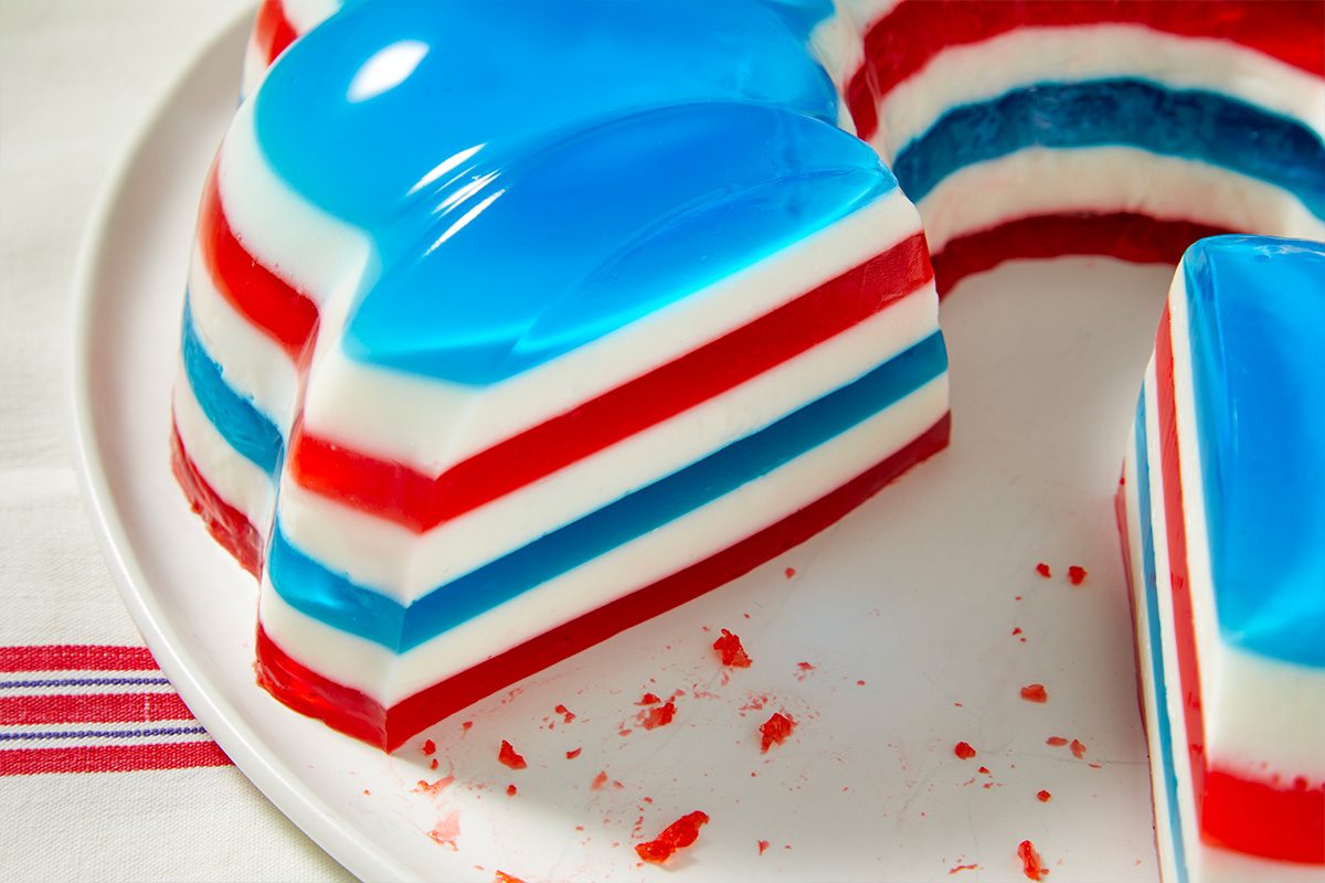 Eye level horizontal image of a festive red, white, and blue layered gelatin dessert served on a white plate. The dessert features distinct stripes of red, white, and blue, with some red gelatin crumbs visible on the plate.