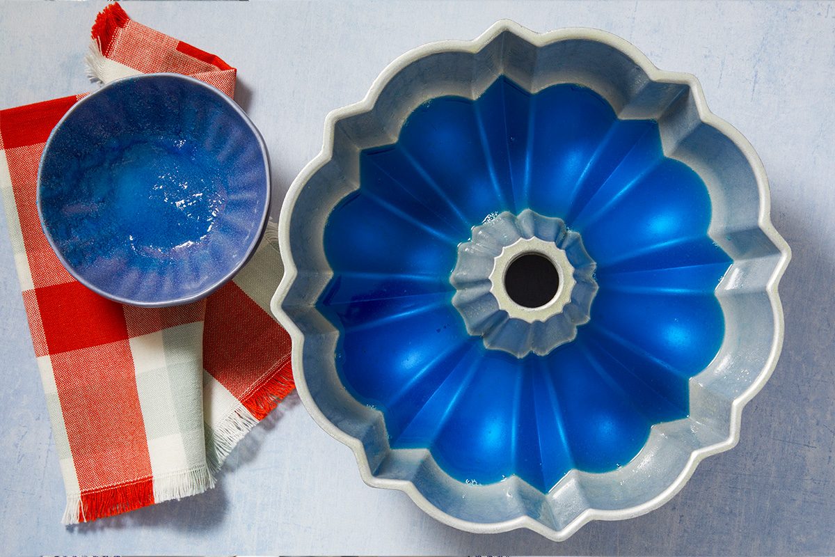 Overhead horizontal image of a silver bundt pan partially filled with vibrant blue gelatin. A small blue bowl with residue of the blue gelatin sits next to a red and white plaid dish towel on a light textured surface.