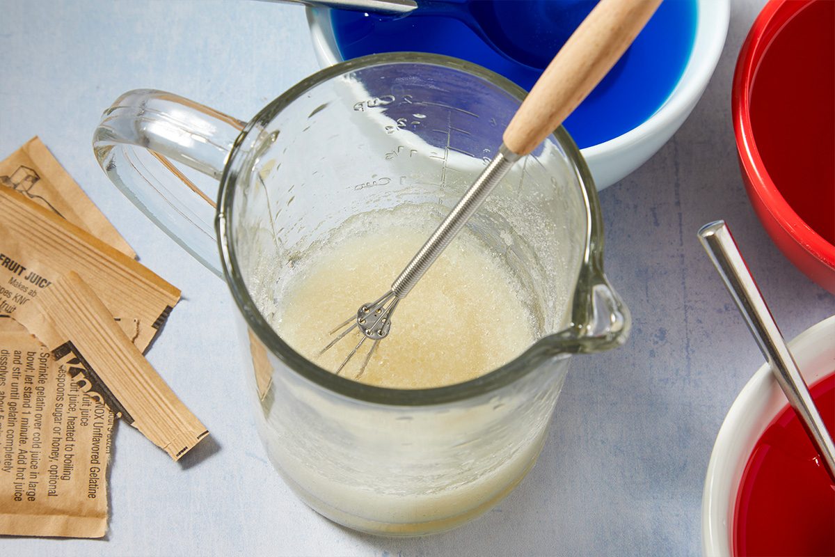 Overhead horizontal image of a glass measuring cup containing bloomed unflavored gelatin with a whisk inside, ready for mixing. Several packets of Knox Unflavored Gelatine are visible to the left, while bowls of blue and red colored liquid are partially visible to the right, indicating a multi-colored gelatin recipe.