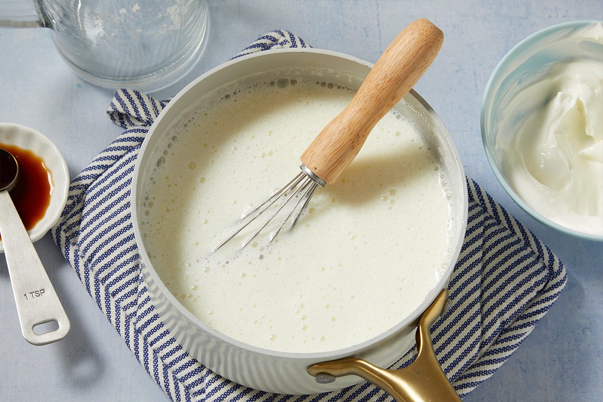 Overhead horizontal image of a light-colored, foamy mixture in a saucepan with a whisk resting inside. Nearby, a small bowl holds dark vanilla extract with a "1 TSP" measuring spoon, and a light blue bowl contains creamy white yogurt. A striped blue and white cloth is positioned beneath the items.