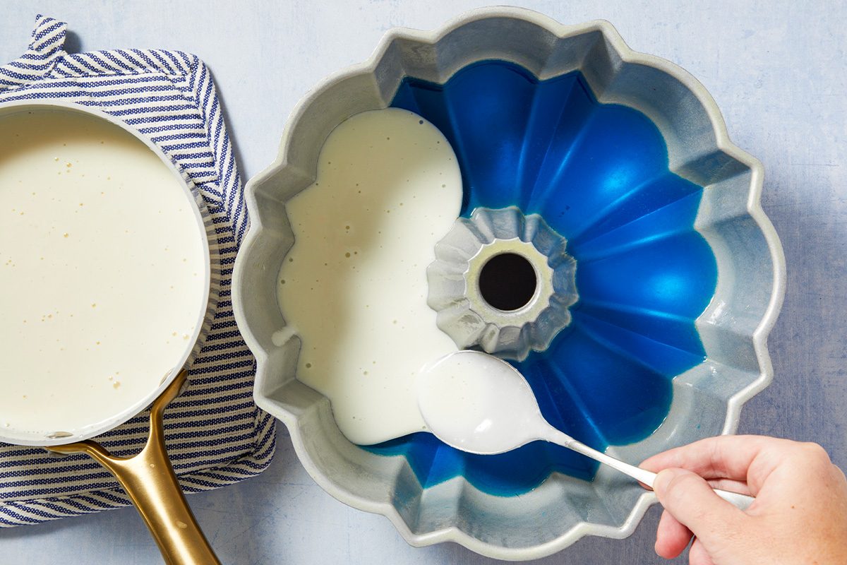 Overhead horizontal image of a hand using a white spoon to spread a creamy, off-white mixture into a blue-lined bundt pan. A saucepan filled with the same creamy mixture rests on a striped towel beside it.