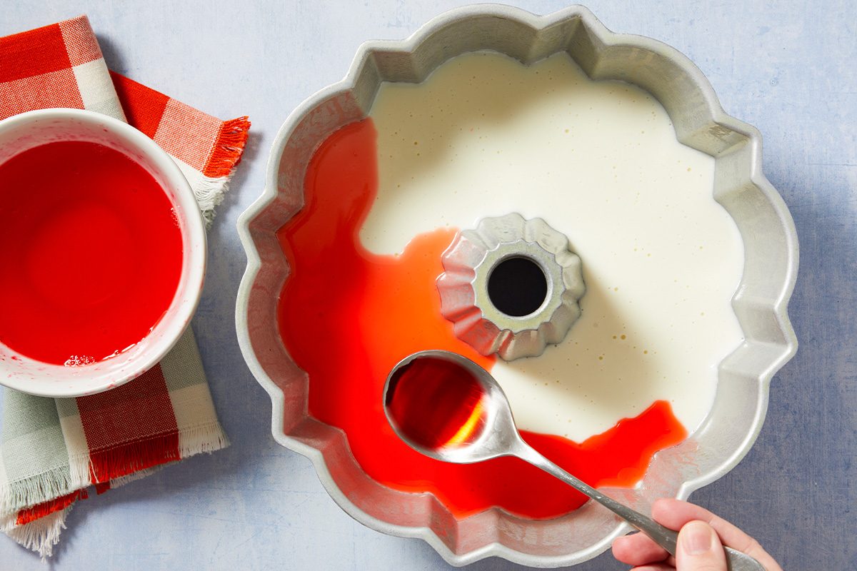 Overhead horizontal image of a hand using a spoon to add vibrant red liquid into a fluted bundt pan. The pan already contains a creamy white liquid, with some red liquid forming a distinct layer. A white bowl filled with more red liquid and a red and white checkered napkin are visible on the light blue surface.