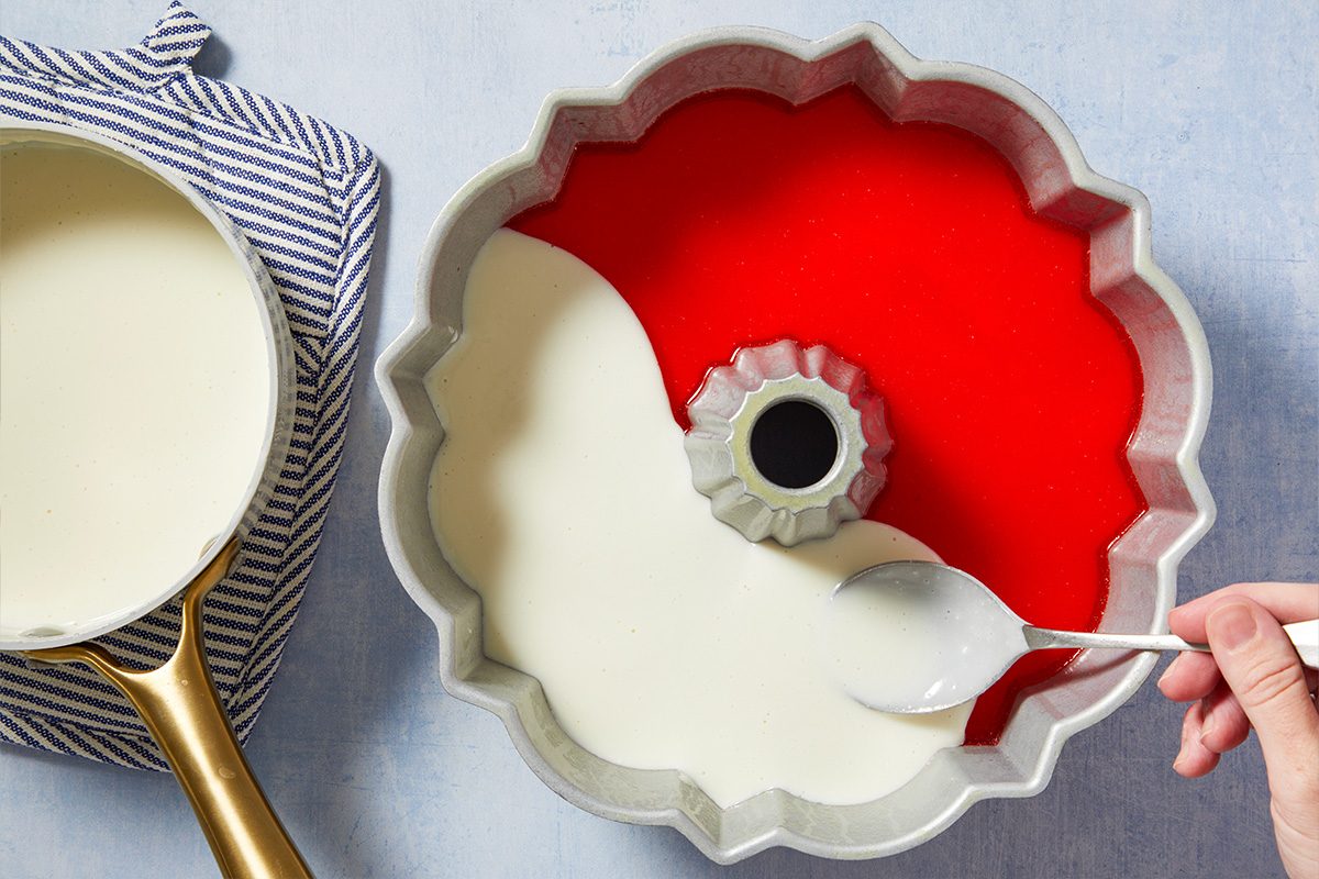 Overhead horizontal image of a hand using a spoon to add a white creamy layer into a decorative Bundt pan. The Bundt pan already contains a red gelatin layer, and a saucepan filled with more white liquid sits on a striped hot pad beside it.