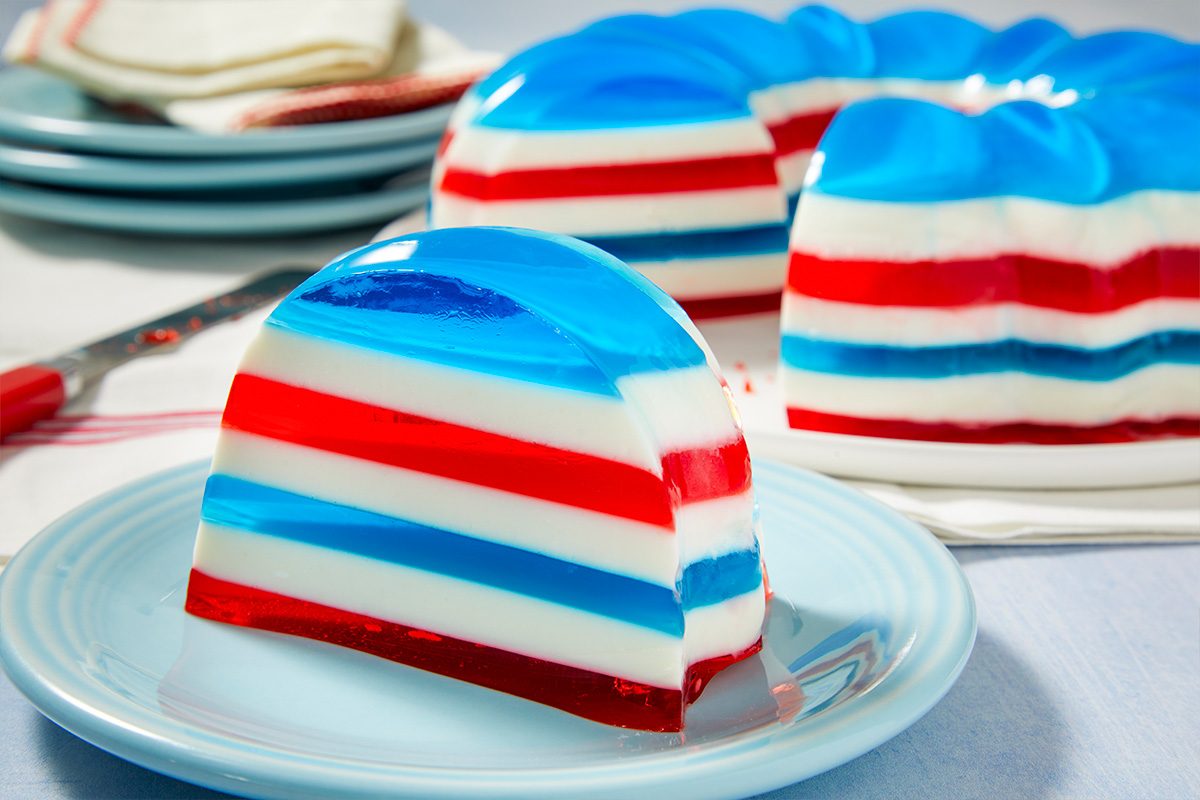 Eye-level horizontal image of a slice of red, white, and blue striped gelatin dessert served on a light blue plate. In the background, the rest of the patriotic gelatin mold is visible. This festive dessert features layers of red, white, and blue gelatin.