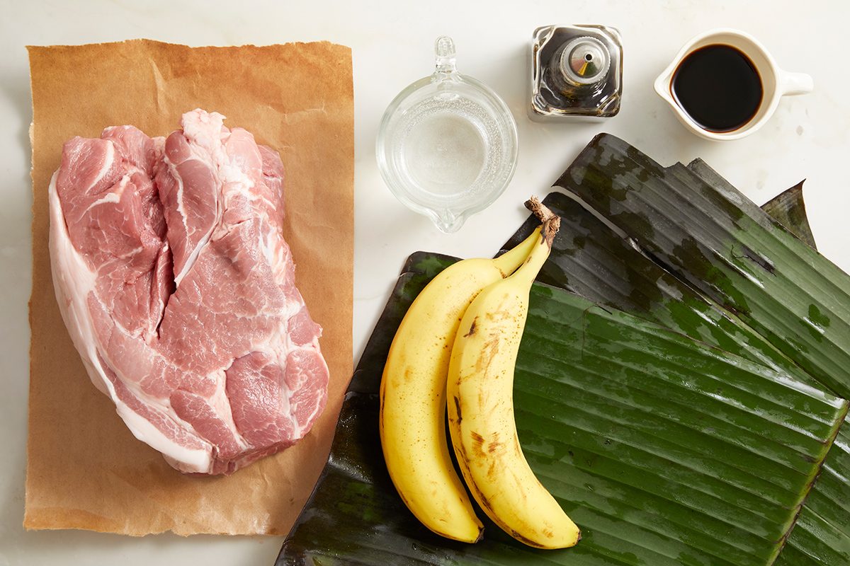 Raw pork on brown paper, two plantains, a glass jug of water, a pepper grinder, a small jug of dark liquid, and large banana leaves arranged on a white surface.