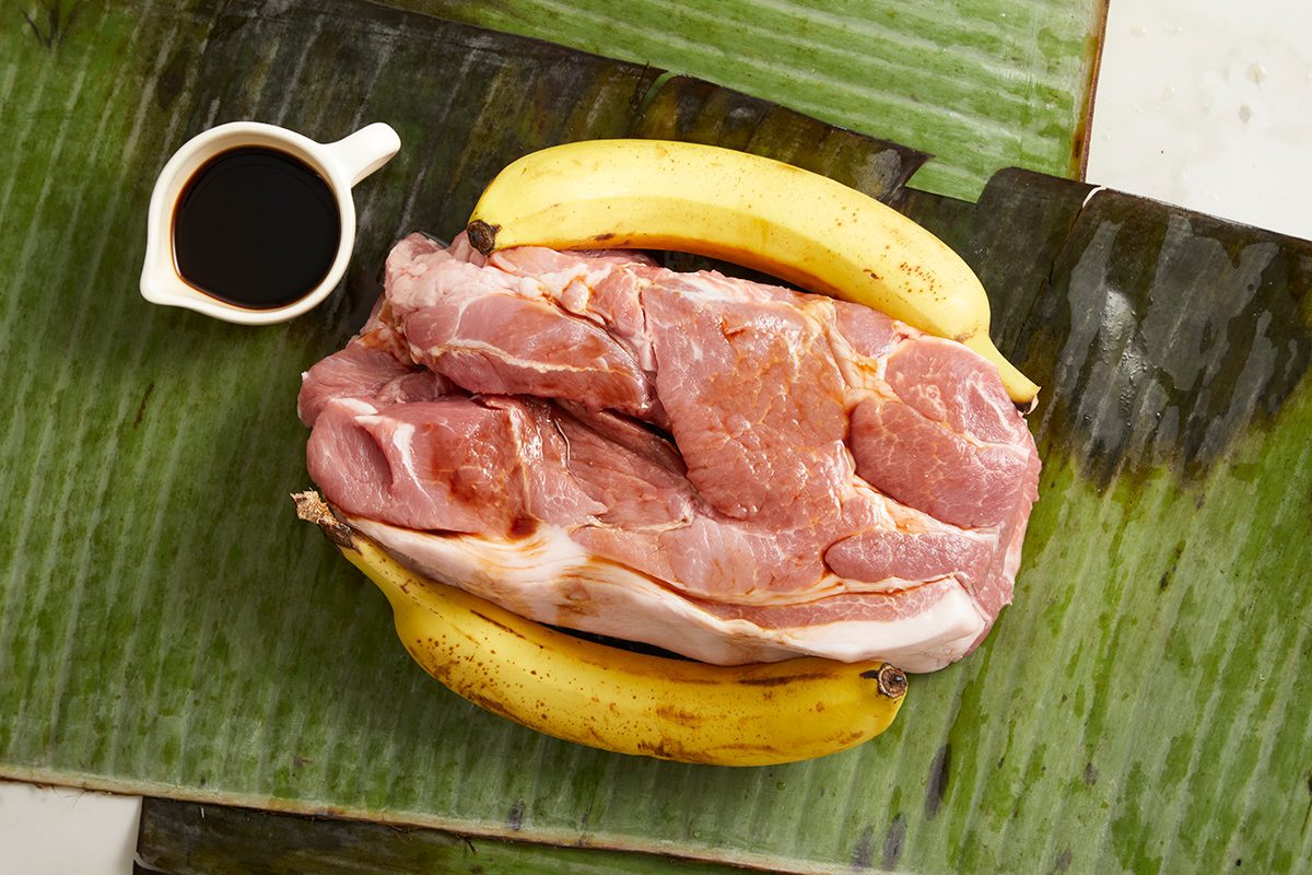 Raw pork sits on banana leaves with two whole yellow bananas beside it and a small white cup of dark sauce on the side.