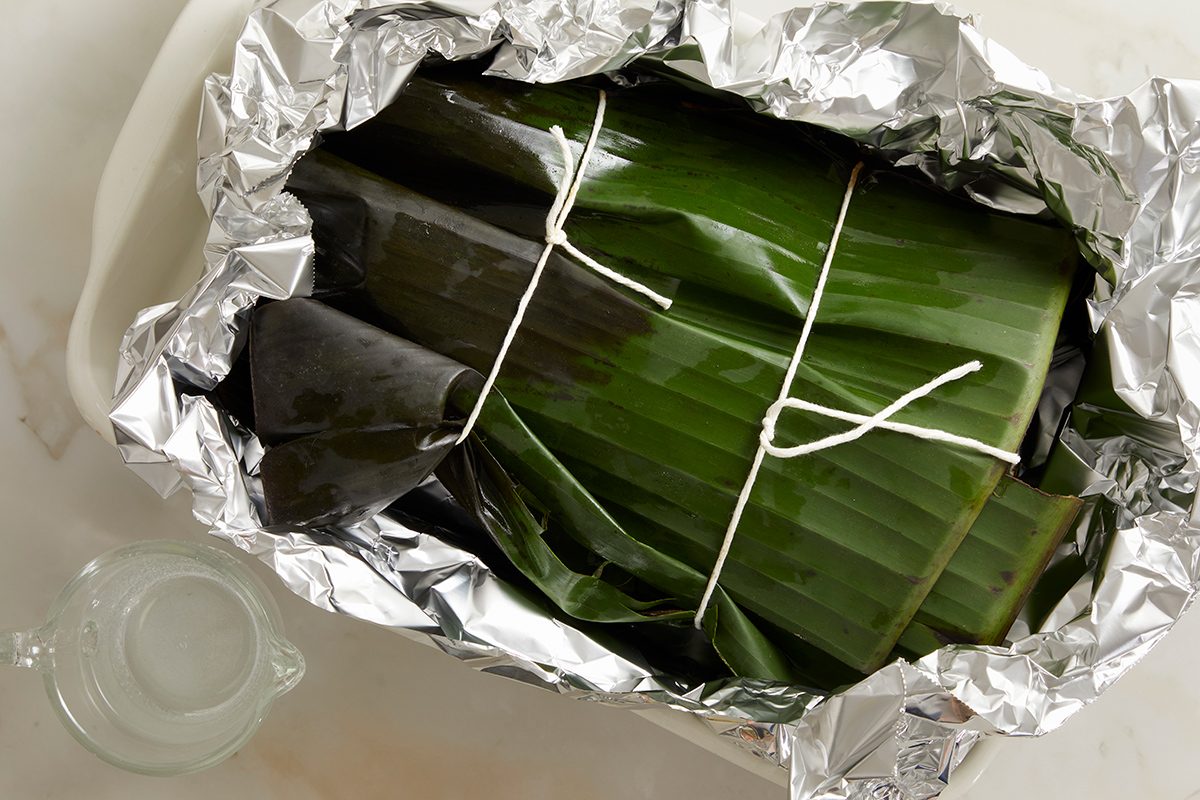 A dish wrapped in green banana leaves and tied with white string, placed inside a tray lined with aluminum foil. A small clear container sits nearby on a light surface.