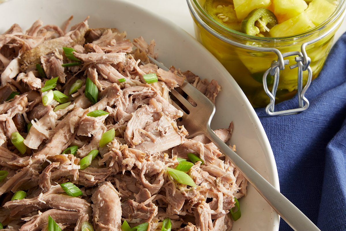 A white dish filled with shredded pork garnished with chopped green onions sits next to a glass jar of sliced pickles and jalapeños on a blue cloth. A metal serving fork rests in the pork.