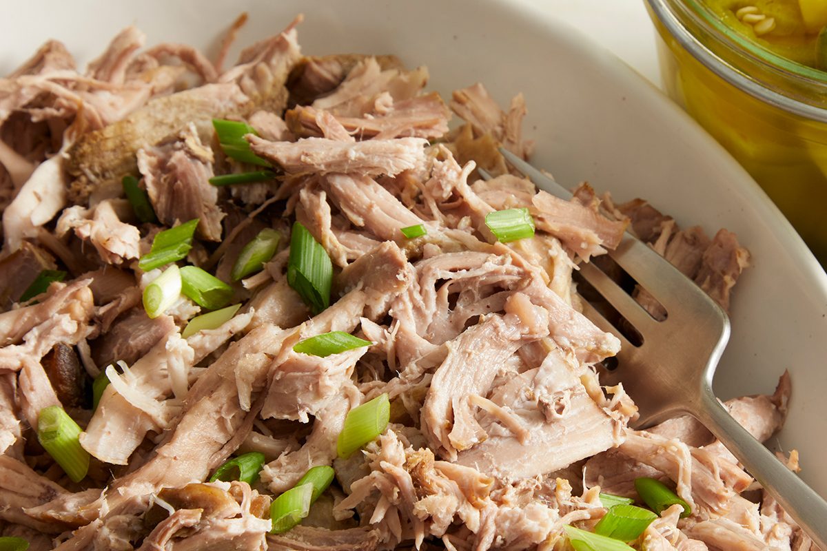 Shredded cooked pork garnished with chopped green onions in a white dish, with a serving fork and a jar of pickles partially visible beside it.