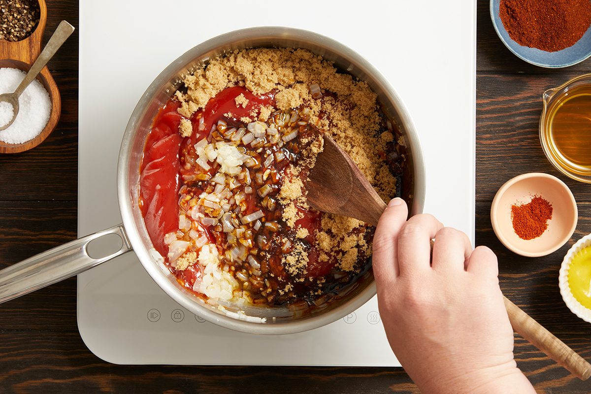 A hand stirs a mixture of chopped onions, ketchup, brown sugar, and spices in a saucepan on a stovetop; small bowls of ingredients are arranged nearby.
