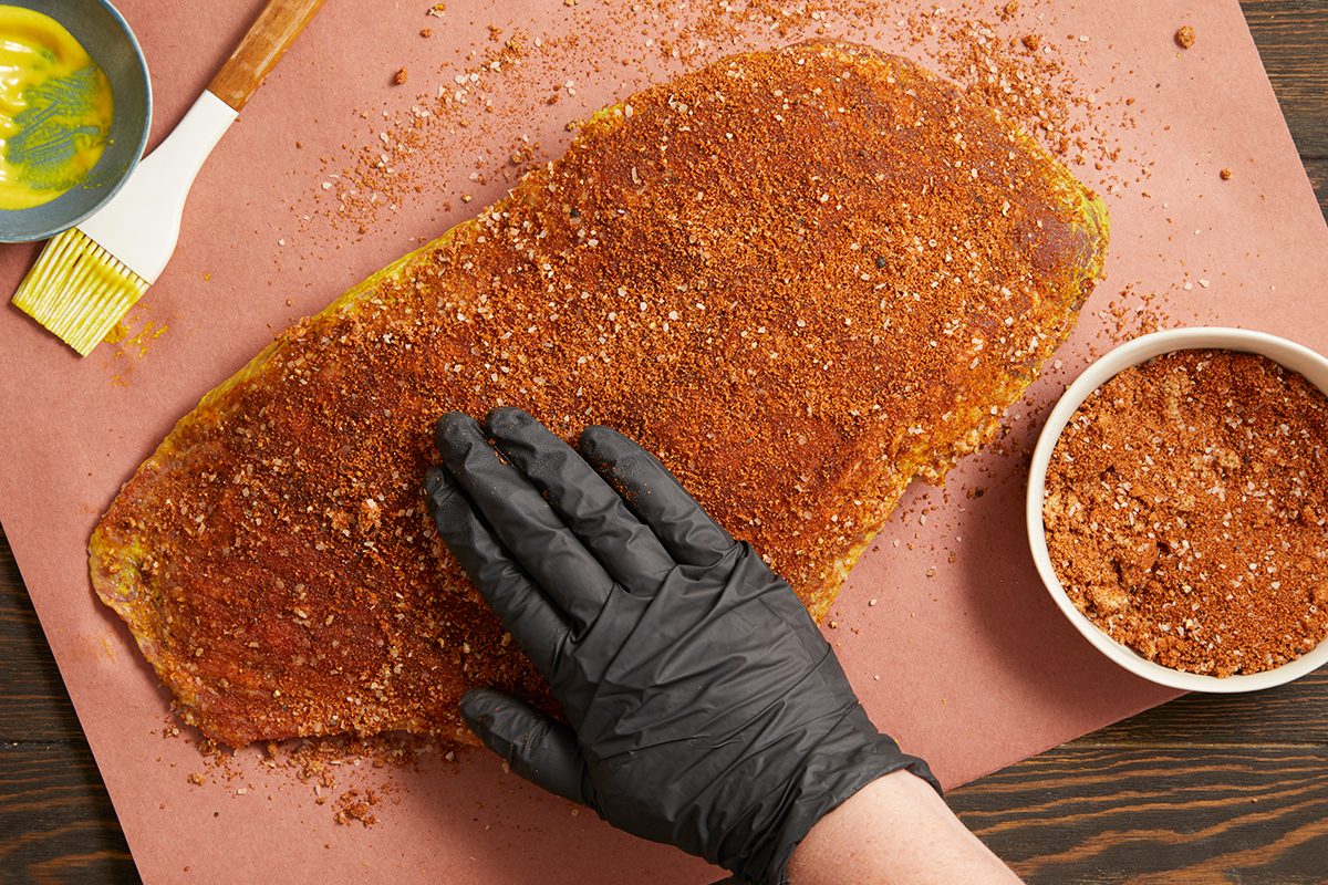 A gloved hand presses a spice rub onto a slab of meat on brown paper, with a bowl of seasoning and a brush coated in yellow marinade nearby.