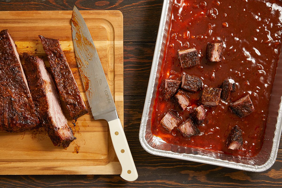 Sliced barbecue ribs on a wooden cutting board with a large knife, next to a tray of sauced burnt ends in barbecue sauce on a dark wooden surface.