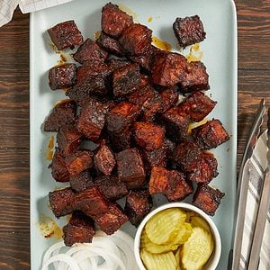 A platter of dark, glazed smoked meat cubes, likely burnt ends, is served with sliced onions and a small bowl of pickle chips on a light rectangular plate. Metal tongs rest on the side.