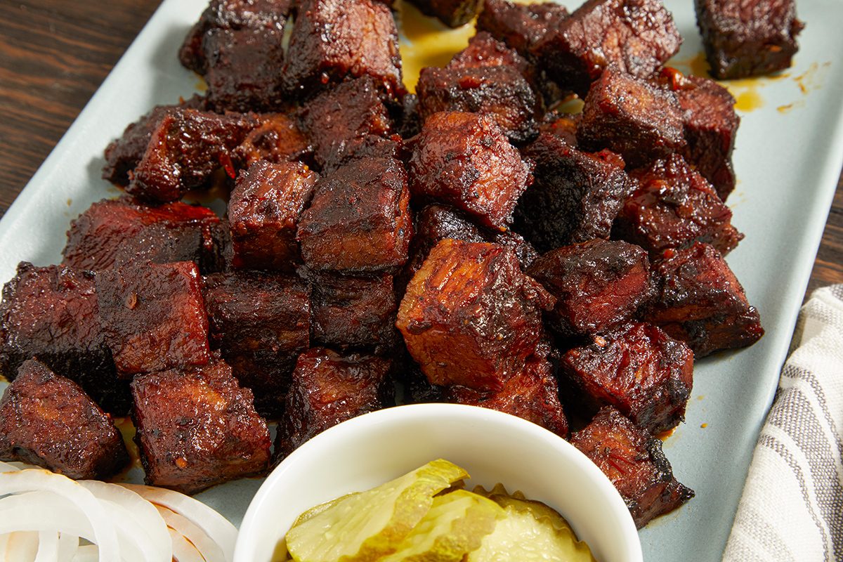 A plate of barbecued burnt ends cut into cubes, served with a side of sliced pickles and onions. The meat appears glazed and caramelized, with a dark, smoky exterior.