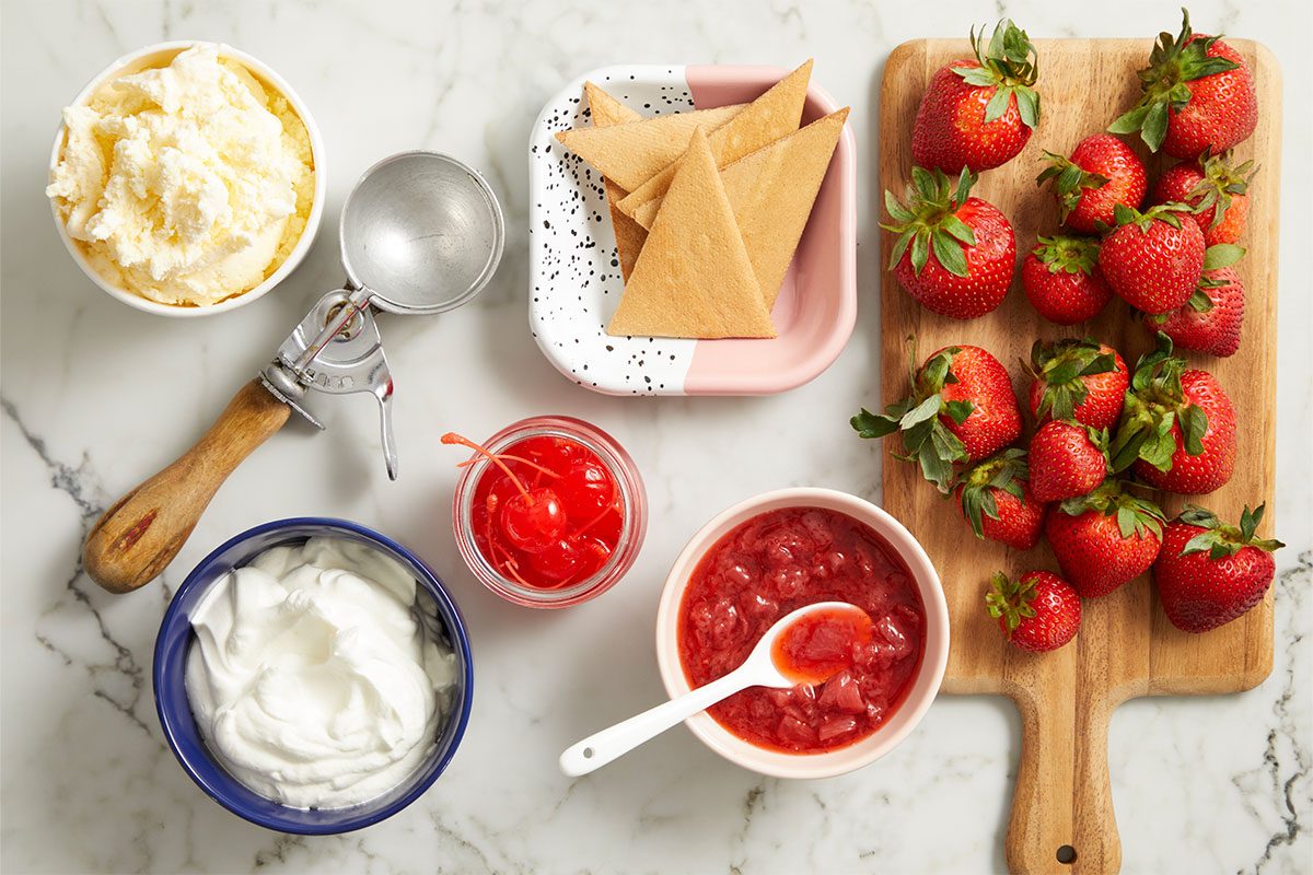 Overhead horizontal image of various dessert toppings and ingredients arranged on a white marble surface. A bowl of vanilla ice cream, an ice cream scoop, whipped cream, maraschino cherries, strawberry topping, fresh strawberries on a wooden board, and triangular wafer cookies are visible.