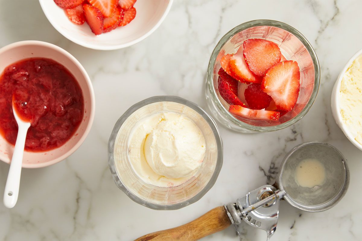 Overhead horizontal image of dessert ingredients arranged on a white marble surface. A scoop of vanilla ice cream sits in one glass, while another glass holds sliced fresh strawberries. Nearby, bowls contain additional sliced strawberries and a red strawberry sauce, next to an ice cream scoop.