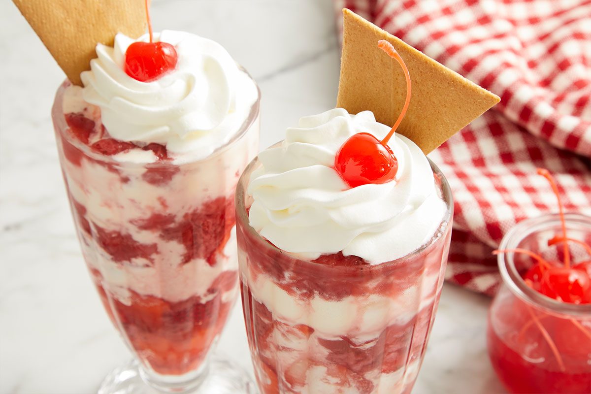Overhead horizontal image of two tall dessert glasses filled with layers of a red fruit swirl and white creamy base, possibly ice cream or parfait. Each dessert is topped with a generous dollop of whipped cream, a bright red maraschino cherry, and a golden rectangular wafer cookie. A small jar of maraschino cherries and a red and white checkered cloth are visible in the background.