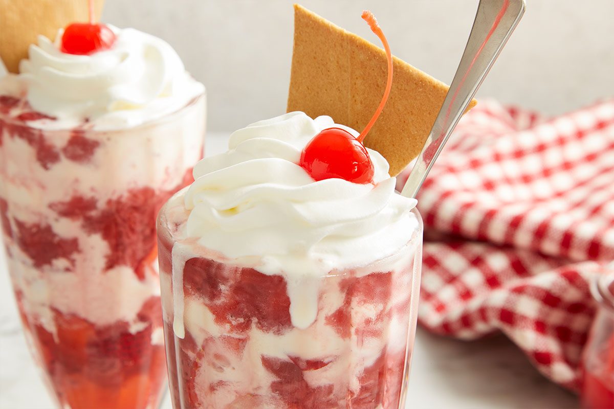 Eye Level horizontal image of two tall glasses filled with a swirled strawberry and vanilla ice cream dessert. The front glass, in sharp focus, is topped with whipped cream, a maraschino cherry, a piece of waffle cookie, and a spoon, with a red and white checkered napkin visible in the background.