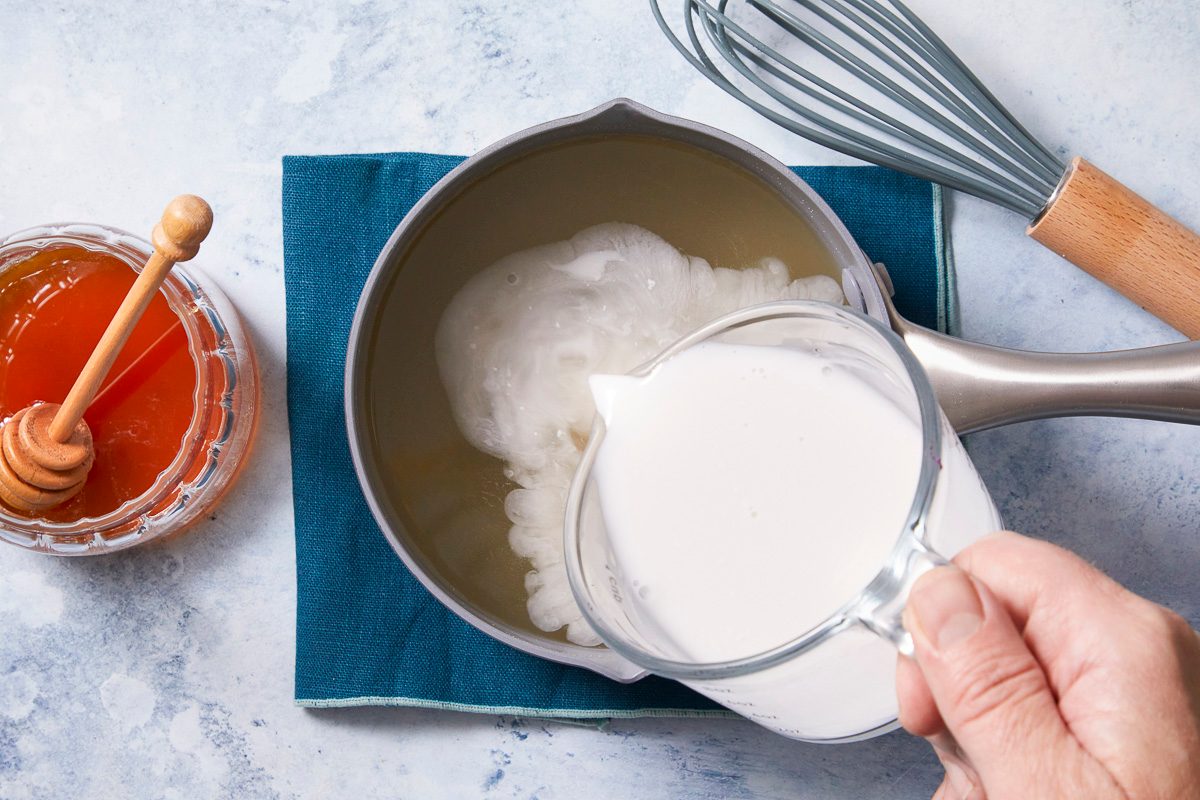 Overhead horizontal image of a hand pouring milk from a glass measuring cup into a saucepan containing a light-colored liquid. A jar of honey with a wooden dipper is visible on the left, and a whisk is in the background.