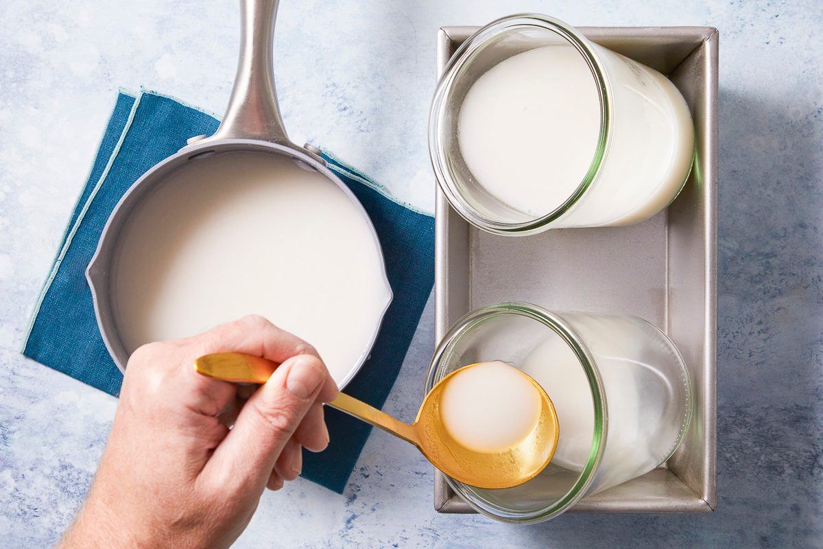 Overhead horizontal image of a hand scooping a white liquid from a saucepan with a golden spoon into a glass jar. Two glass jars filled with the liquid are nested in a metal loaf pan, resting on a blue linen napkin.
