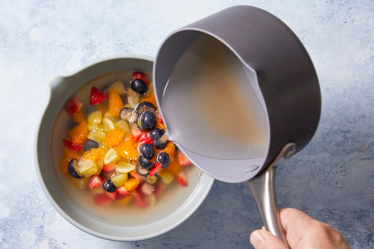 Overhead horizontal image of a hand pouring clear liquid from a gray saucepan into a light gray bowl. The bowl contains chopped strawberries, blueberries, oranges, and green grapes, partially submerged in liquid.