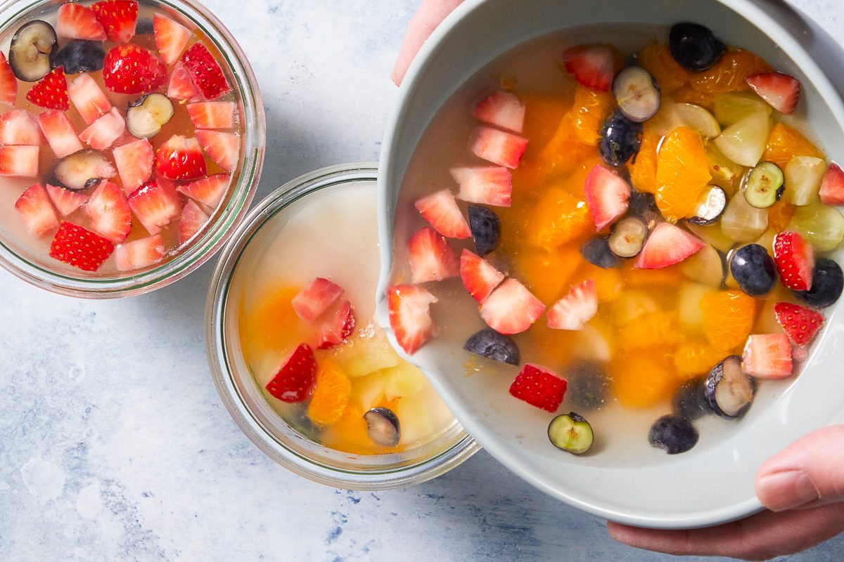 Overhead horizontal image of a hand pouring a colorful mix of fruit and liquid from a white pitcher into a glass jar. The jar is being filled with diced strawberries, blueberries, orange segments, and grapes, while another glass jar on the left is already filled with similar fruit in a light liquid.