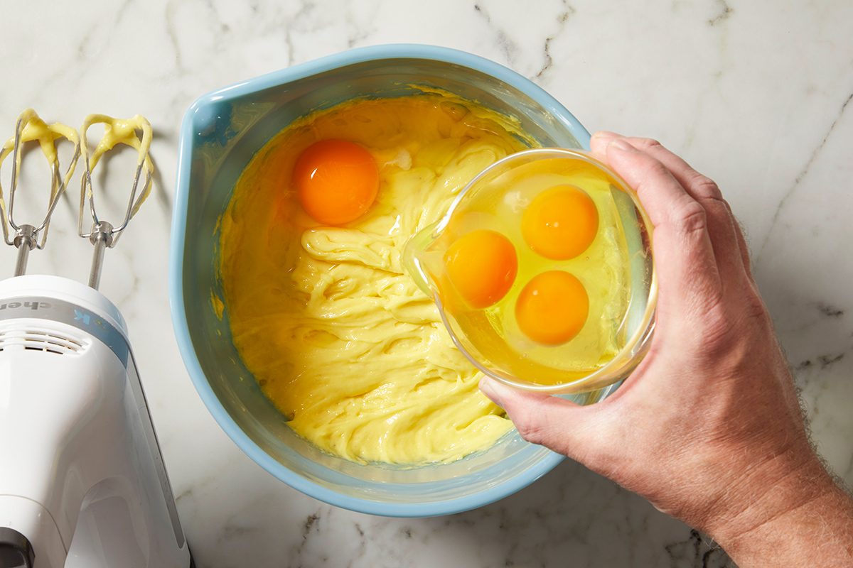 A hand holds a bowl with three eggs above a mixing bowl containing batter and two cracked eggs. An electric hand mixer with batter residue is next to the mixing bowl on a marble countertop.
