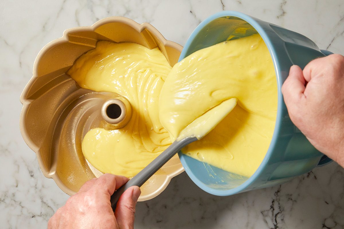A person pours yellow cake batter from a blue mixing bowl into a bundt pan on a marble countertop, using a spatula to help guide the batter.