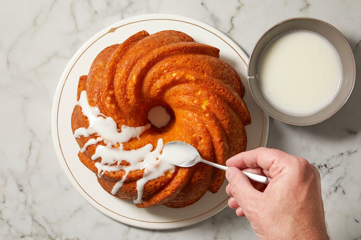 A hand is drizzling white icing onto a Bundt cake with a spoon. The cake sits on a white plate, and a bowl of extra icing is nearby on a marble surface.