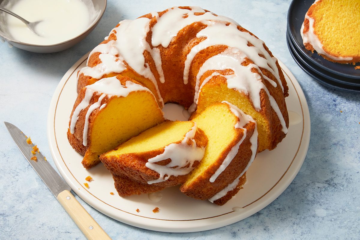 A round bundt cake with white icing drizzled on top sits on a plate. A couple of slices are cut, revealing the golden yellow inside. A knife and dark plates are nearby on a light surface.