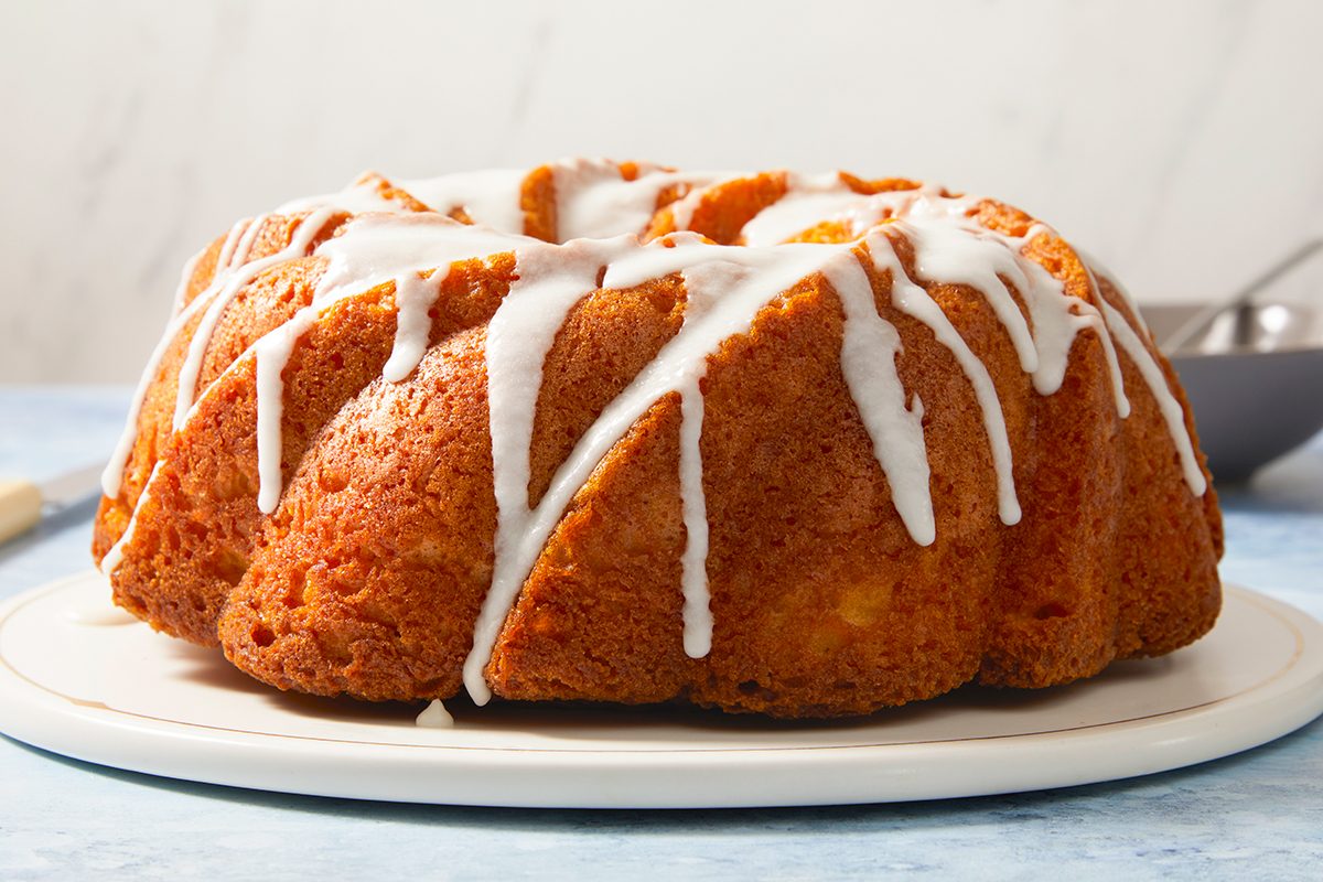 A golden brown bundt cake is displayed on a white plate, topped with a drizzle of white icing. The background is light and simple, drawing focus to the cake.