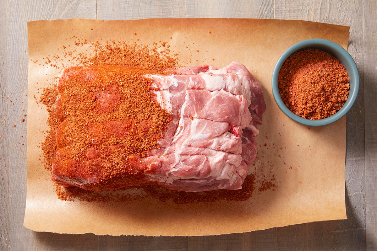 A raw pork roast sits on parchment paper, partially covered with a reddish-brown dry rub seasoning. A small blue bowl filled with more dry rub is placed nearby on the paper.