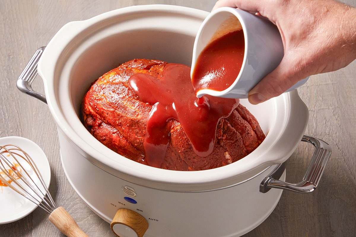 A person pours red sauce from a small pitcher over a large piece of meat in a white slow cooker. A whisk and small plate are beside the cooker on a gray surface.